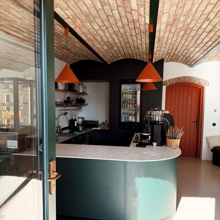 Zelený barový pult s terrazzovou deskou pod cihlovým klenutým stropem a oranžovými lampami
